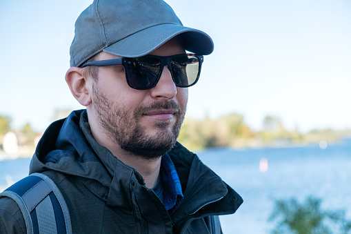Bearded man in sunglasses and a baseball cap against the backdrop of a lake or river on a sunny autumn day. Concept of outdoor recreation, solitude or hiking