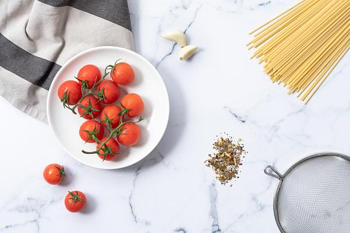 Flat lay view of cherry tomatoes, spaghetti, garlic, herbs and colander on a marble table.