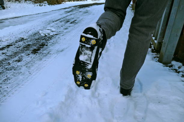 左足を持ち上げる人の黒いブーツの裏には、スリッププロテクションとスパイクが付属しています。黒い氷と雪の中で冬の靴のための安全装置。 - スパイクシューズ ストックフォトと画像