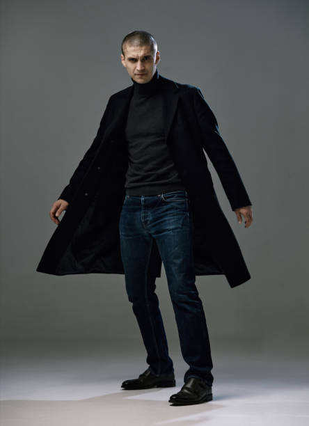 istock Confident serious handsome guy in black coat, detective or security on gray studio background, full-length male portrait 1299644364