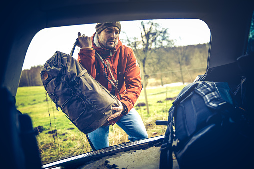 Male hiker with backpack, knitted hat and hip red jacket, unpacking trunk of his car - ready for a hiking adventure.