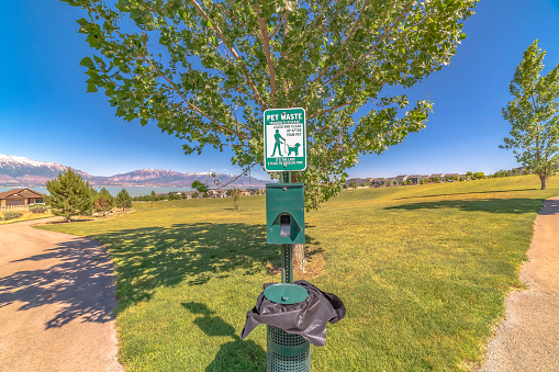 Dog poop bags and garbage can against timpanogos mountains and blue lake. Pathways on vast grassy land with view of houses and blue sky can also be seen in this sunlit landscape.