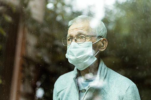Senior man wearing protective, medical mask looking through window, quarantine, during pandemic