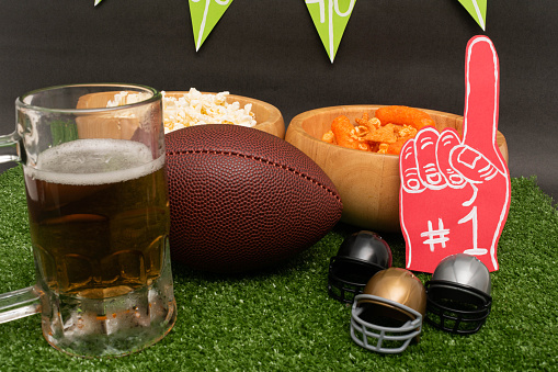 American football party. American football ball, beer, little helmets, snacks and finger foam on green grass and black background