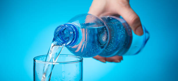 a woman's hand fills a glass with water from a bottle on a light blue background - water water bottle glass pouring imagens e fotografias de stock