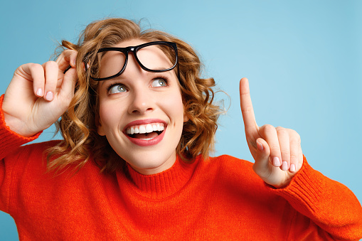 Happy young female lifting stylish glasses and smiling while pointing up against blue background