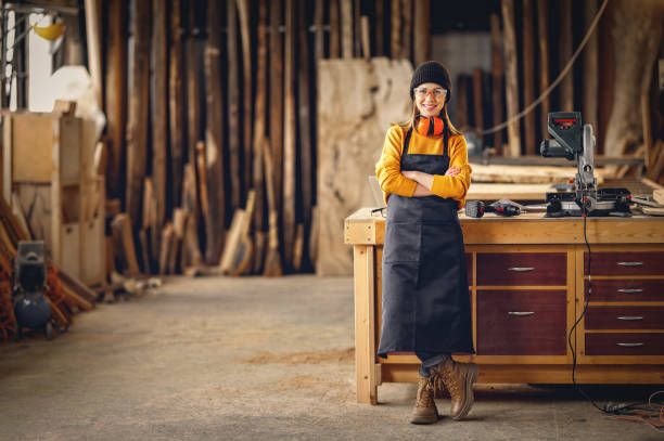 Positive woman working in joinery workshop Full body of smiling confident young female joiner in apron standing near workbench and looking at camera friendly while working in craft workshop carpenter stock pictures, royalty-free photos & images