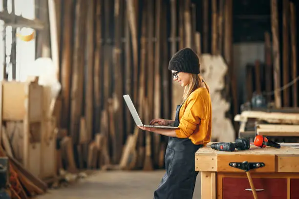 young female carpenter looks drawings on a laptop during  while  break in working with wood in modern handicraft studio