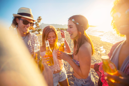 Group of friends drinking beer and dancing on the beach at sunset. They are lifting their bottles in a toast. All are happy and smiling and laughing.