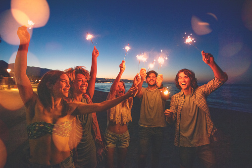 Group of friends playing with sparklers and fireworks on the beach at sunset. They are dancing and having fun . All are happy and smiling and laughing.