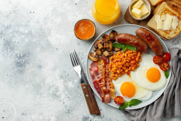 istock Full English breakfast on a plate with fried eggs, sausages, bacon, beans, toasts and coffee on light stone background. With copy space. Top view 1299672604