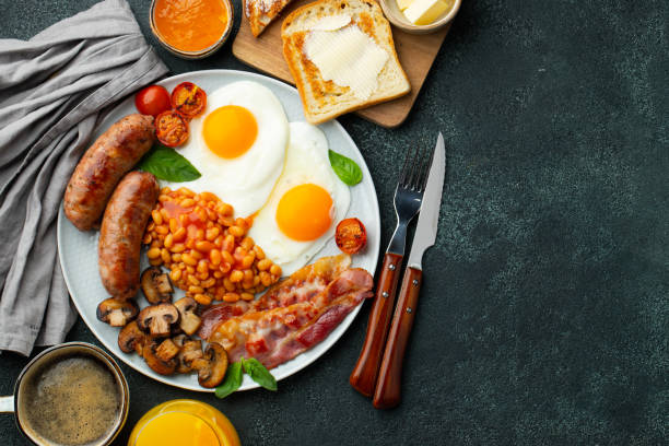 istock Full English breakfast on a plate with fried eggs, sausages, bacon, beans, toasts and coffee on dark stone background. With copy space. Top view 1299672607
