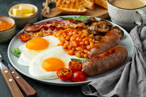 Full English breakfast on a plate with fried eggs, sausages, bacon, beans, toasts and coffee on dark stone background.