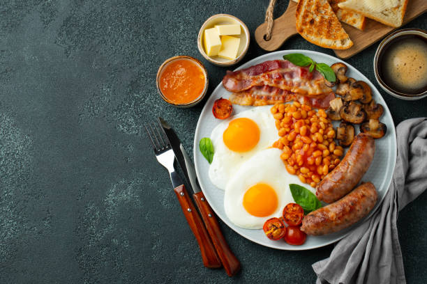 istock Full English breakfast on a plate with fried eggs, sausages, bacon, beans, toasts and coffee on dark stone background. With copy space. Top view 1299672617