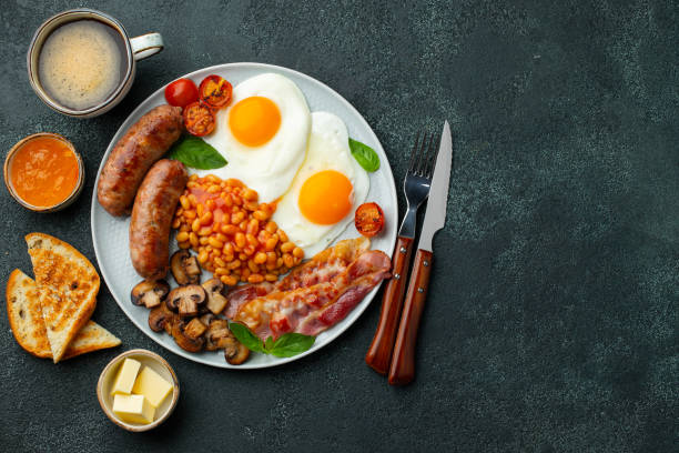 istock Full English breakfast on a plate with fried eggs, sausages, bacon, beans, toasts and coffee on dark stone background. With copy space. Top view 1299672619