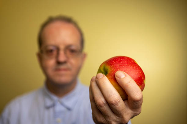 istock A middle-aged man against yellow background, presenting an apple as healthy eating habit with focus on the foreground 1299673614