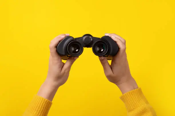 binoculars in hand over yellow background, find and search concept.