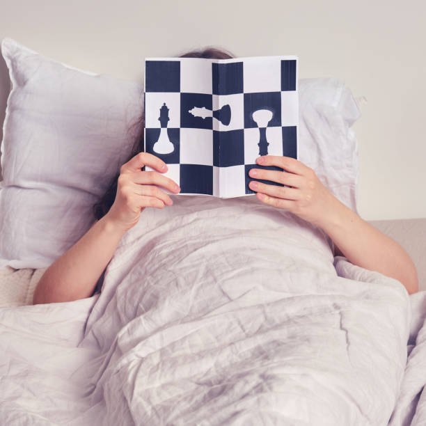 une femme est couchée sur le lit et lit un livre d’échecs. le concept de problèmes dans la vie des gens intelligents - chess board room business strategy photos et images de collection