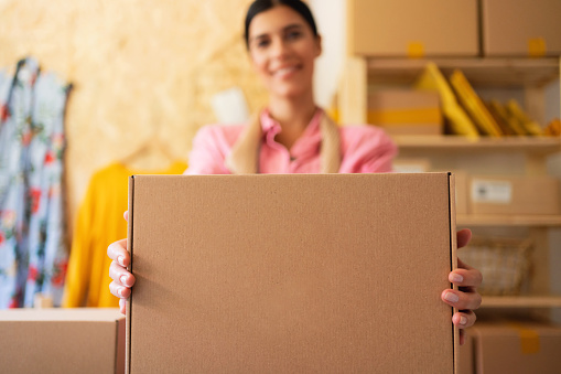 Young female small business owner standing in her home office, holding package ready for home delivery and looking at camera. Online selling. Online shopping. Focus on a cardboard box.