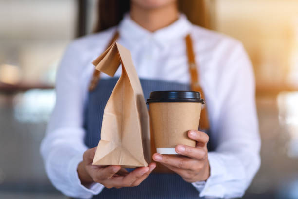 a waitress holding and serving paper cup of coffee and takeaway food in paper bag to customer in a shop Closeup of a waitress holding and serving paper cup of coffee and takeaway food in paper bag to customer in a shop junk food stock pictures, royalty-free photos & images