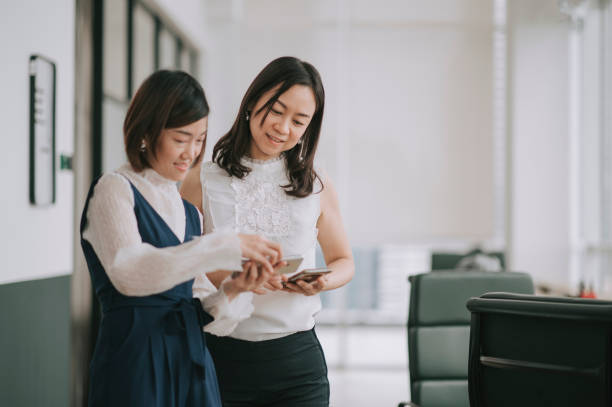 istock 2 asian chinese female white collar workers looking at smart phone talking during break time in office 1299703391