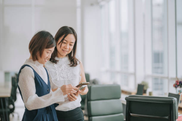 istock 2 asian chinese female white collar workers looking at smart phone talking during break time in office 1299703410