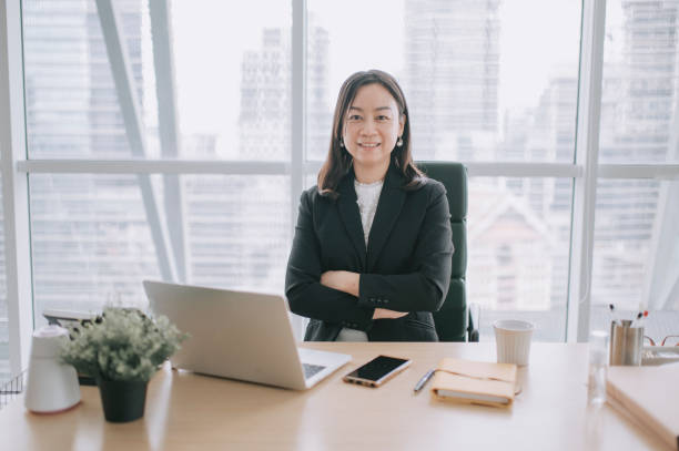 アジアの中国の美しい女性ceoは、オフィスで彼女の机に微笑むカメラを見て - senior adult women adult tan ストックフォトと画像