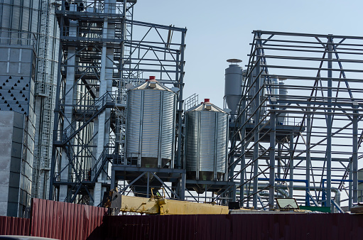 Construction of a modern grain terminal on a blue sky background. Metal cylindrical silo and support metal structure for the loading conveyor. Agribusiness. Export of grain.