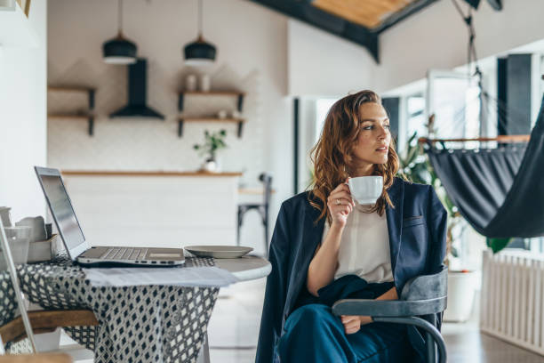 a woman is sitting in the kitchen, distracted from working with her laptop, drinking from a mug and looking thoughtfully away - women professor mature adult human face imagens e fotografias de stock