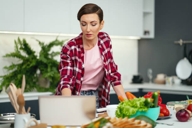 istock Making a video call using laptop computer housewife showing her new house and new kitchen while cooking or baking holding eggs in a hand. Young woman cooking in the kitchen. New house concept 1299722221