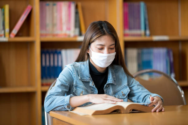 istock Female college student wearing a mask is reading at the university library. 1299728791