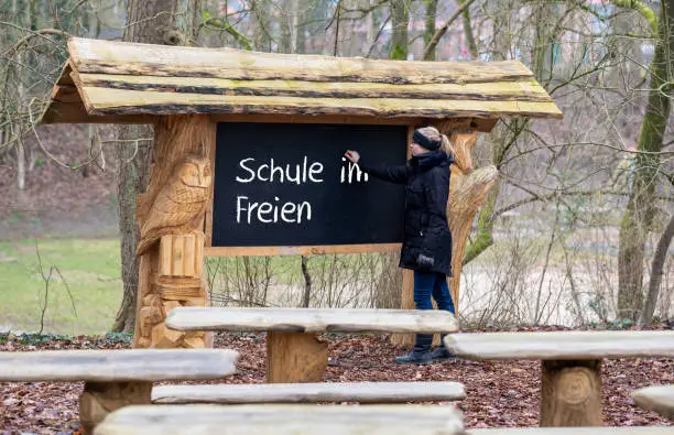 Female teacher writes on a blackboard the German expression "Schule im Freien"(school outside). A classroom in the forest is qualified for teaching during the corona pandemic.