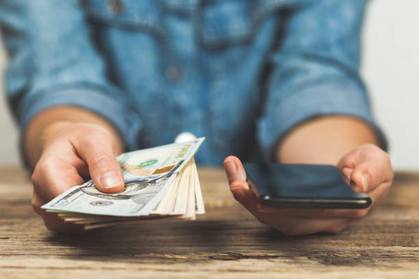 в руках женщина держит доллары и телефон. онлайн денежный перевод - bill mobile phone smart phone currency стоковые фото и изображения