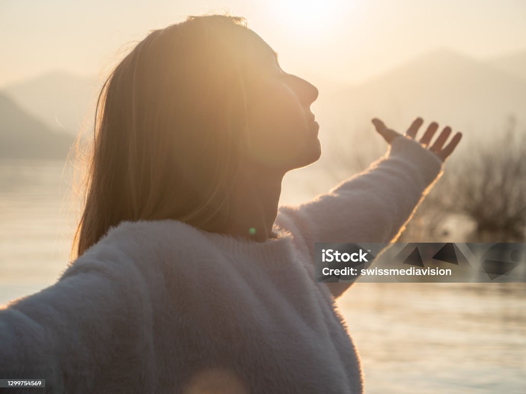 Woman embraces nature, she stands arms out by the lake at sunset She stands arms raised up, closed eyes, mountains and lake in distance Gratitude Stock Photo