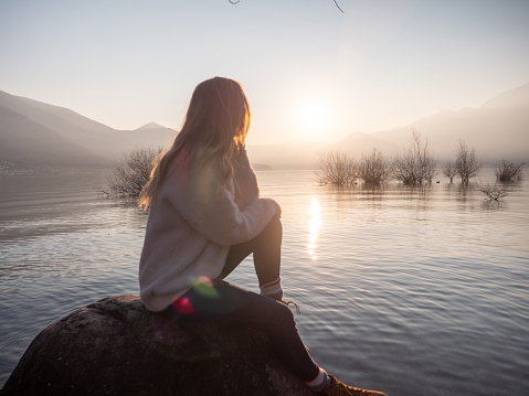 She sits on a big rock at the edge of the mountain lake, sun setting in distance