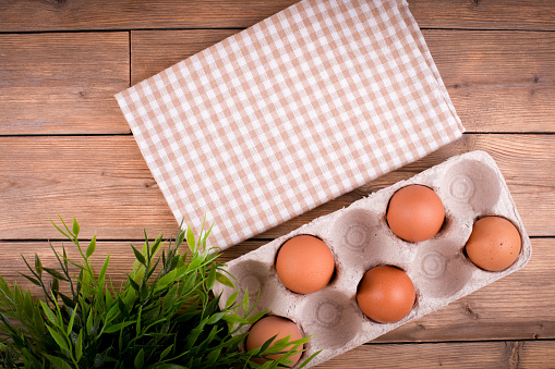 Close-up of raw chicken eggs in an egg box on a wooden background, a flower in a pot, a towel in a cage. Copy space