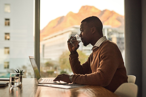 Shot of a young businessman drinking water while working on a laptop in an office