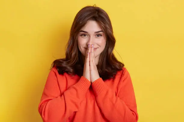 Pretty Caucasian woman keeps palm together, has pleased expression, asks for something, looks satisfied, wearing orange casual jumper, standing isolated over yellow background.