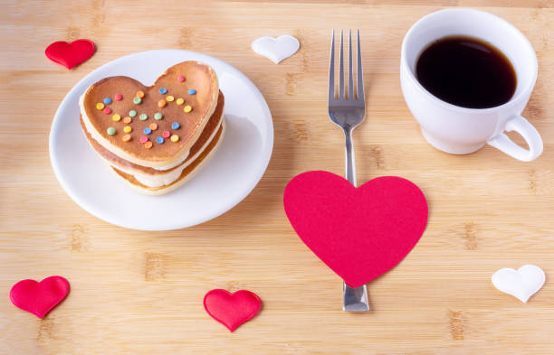 istock Homemade heart shaped pancakes with sugar decoration, a fork on a red heart shape, a mug with coffee or cocoa 1299767501