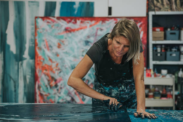 Young woman paints an abstract picture with her hands and spatula on a work table in her interior studio. Focus on head stock photo