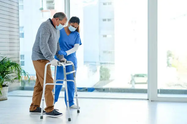 Shot of a masked senior man using a walker with the assistance of a nurse