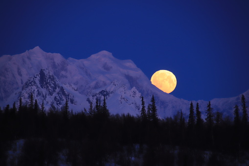The full moon sets behind the mountains of the Alaska Range near Denali on a cold winter morning.