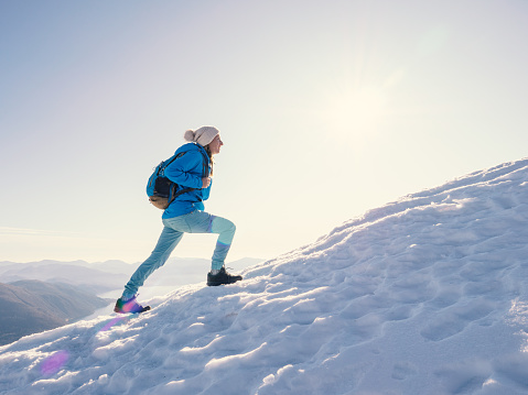 Woman hiker walking uphill on snowy mountain ridge. Sportive young female on a hike ascending mountain in winter. Relentless achievement concept