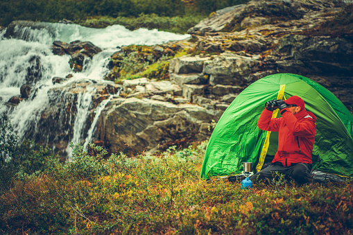 Caucasian Outdoor Men in Front of His Tent Spotting Something Using Binoculars. Scenic Landscape with Waterfall in Background. Wilderness Camping Spot.