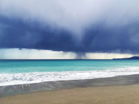 View of a storm on the blue emerald sea, horizon on water and shore with white surf on the waves, cloudy dark sky with rain in the background