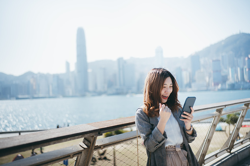 Asian young woman using her smartphone by the promenade, she looks happy as she has got a good news from her job application.