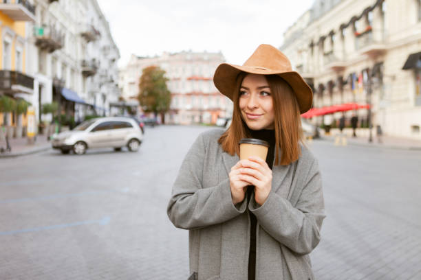 istock Stylish millennial woman in a slicker coat and felt hat jerks a coffee cup on the go while walking in a european city 1299804004