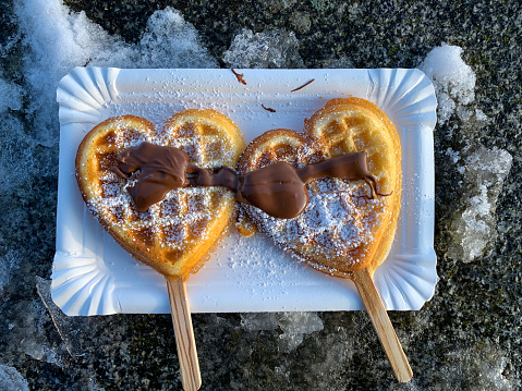Two heart shaped waffles on a sticks with chocolate and powdered sugar on paper plate on frozen snowy surface directly above view