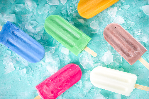 Selection of bright multicolored ice cream popsicle. Various gelato, frozen lollypops - chocolate vanilla blueberry strawberry pistachio orange, with crushed ice on light blue sunny background