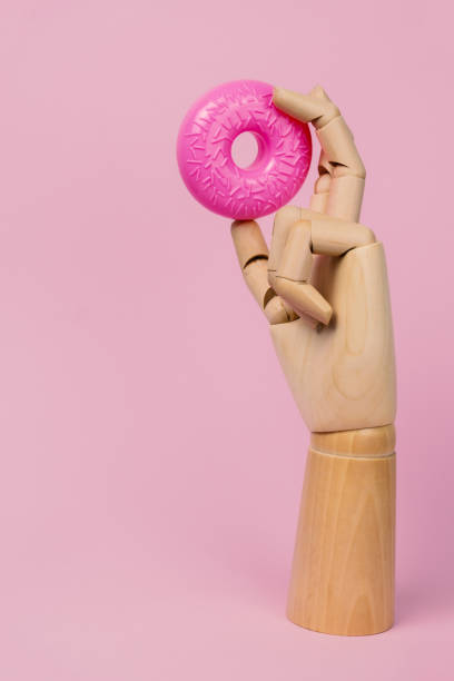 istock Wooden hand with pink donut on a pastel pink background. 1299809598